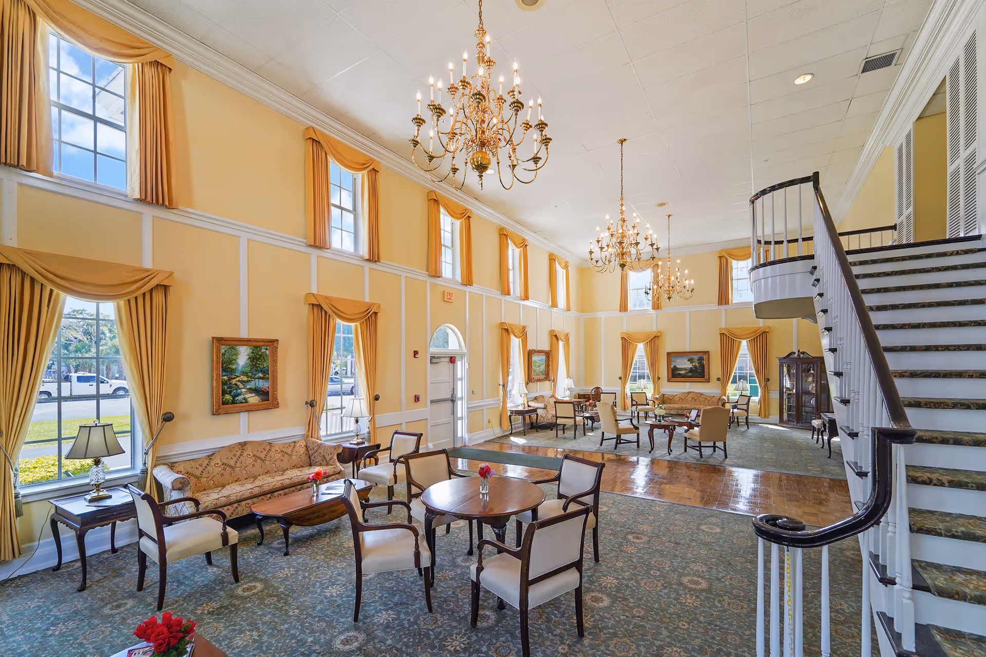 A spacious and elegant living room area with high ceilings, large windows with yellow curtains, multiple chandeliers, and classic furniture including sofas, armchairs, and round tables. A staircase with a dark wooden handrail is visible on the right side.