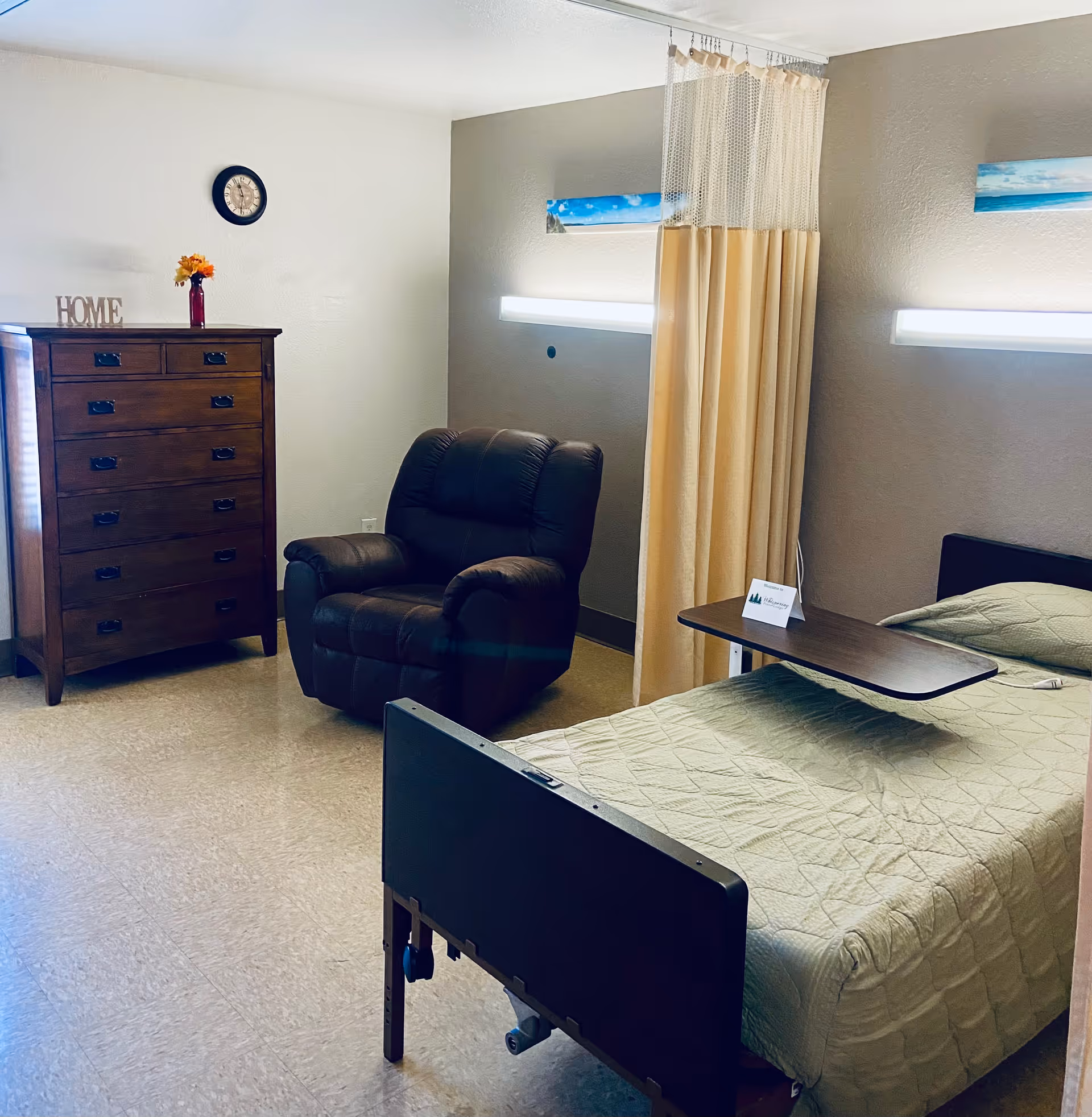A senior living facility bedroom with a single bed covered in a light green quilt, a brown recliner chair, a wooden chest of drawers with a small vase of flowers and a decorative 'HOME' sign on top, a wall clock, and a privacy curtain partially drawn around the bed.