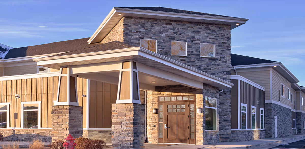 Exterior view of a modern assisted living facility building with stone and wood paneling, a covered entrance supported by stone pillars, and a wooden door with glass panels. The sky is clear and blue.