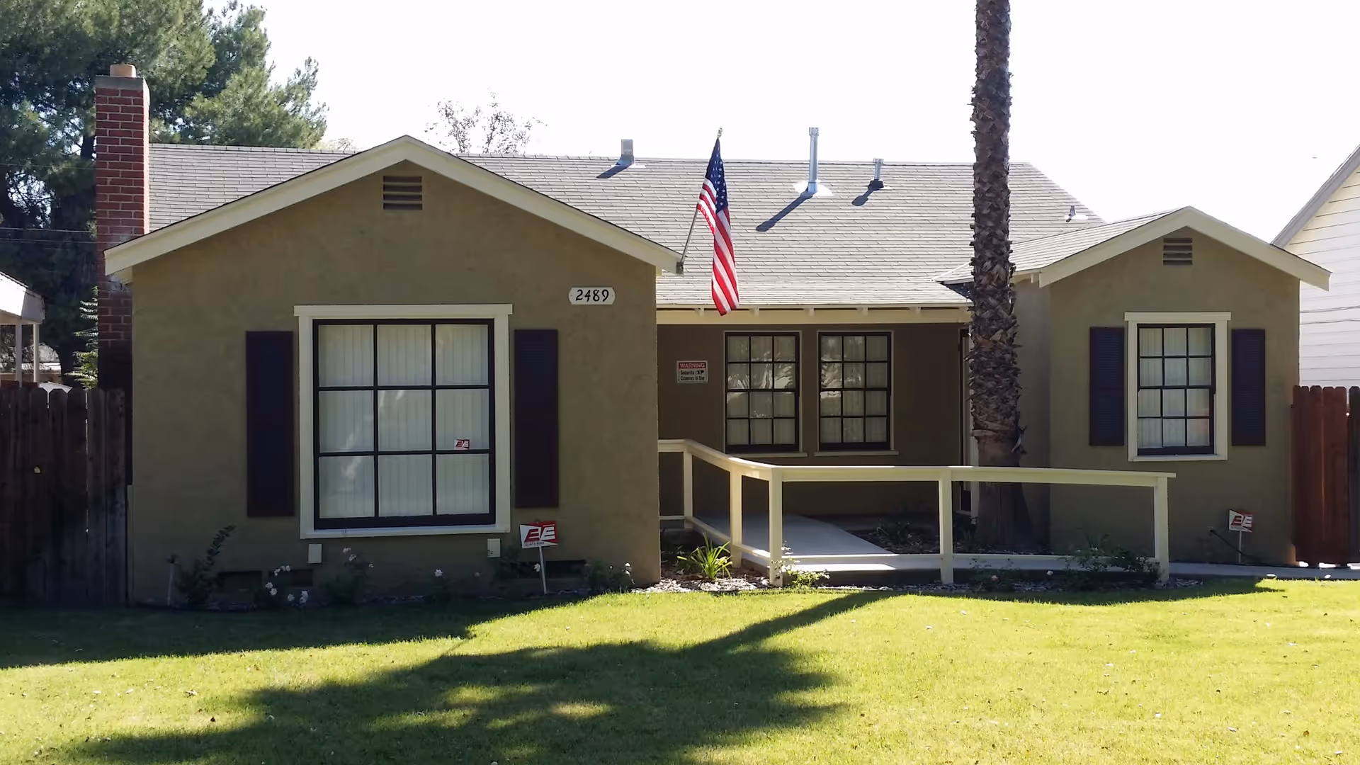 Single-story house with beige exterior, three front windows with dark shutters, a chimney on the left side, an American flag hanging near the center, and a wheelchair ramp leading to the front door. The house number 2489 is visible above the left window. The lawn in front is green and well-maintained with a tall palm tree near the center.