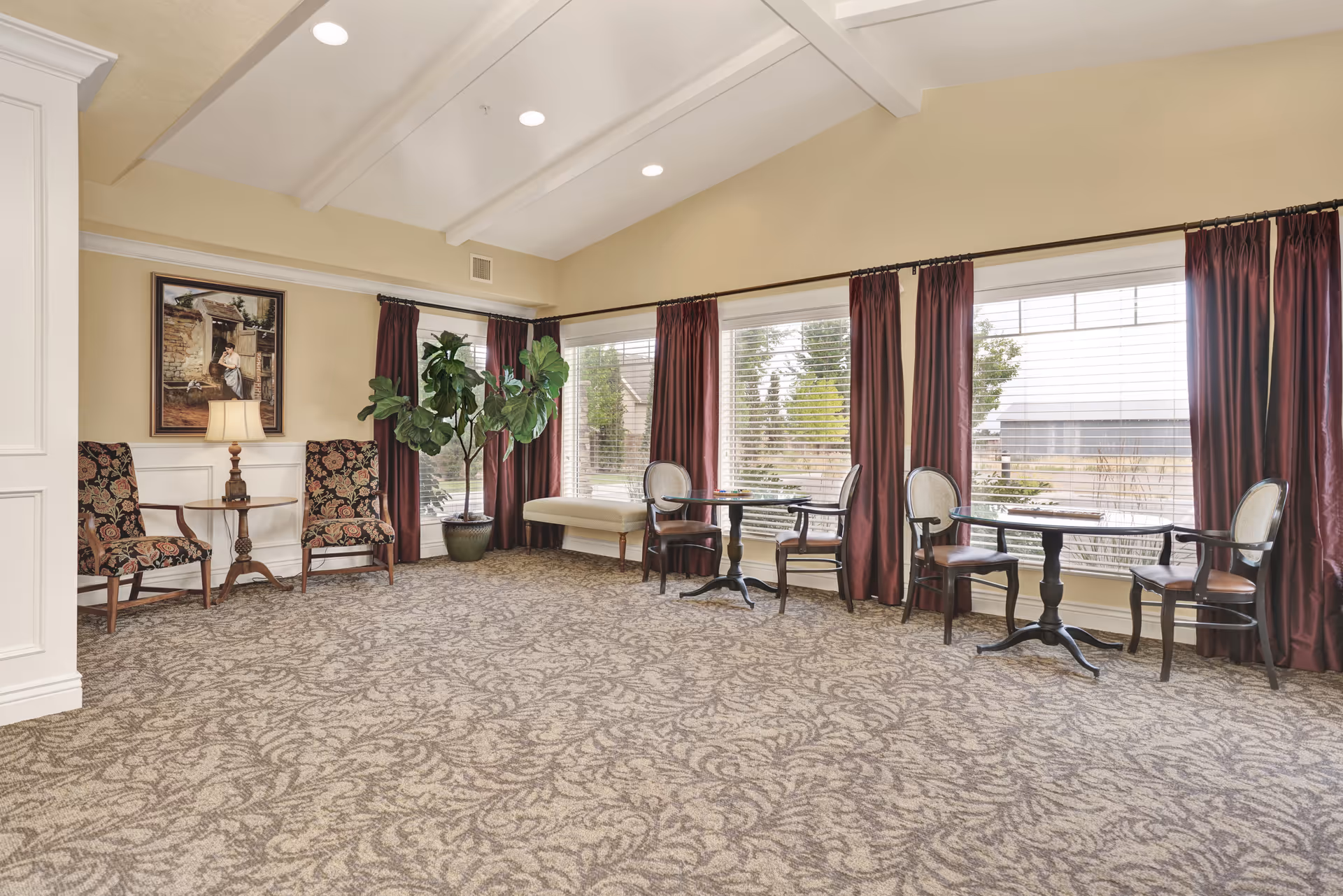 A bright and spacious sitting area with large windows covered by maroon curtains. The room features patterned carpet, two small round tables each with two chairs, a bench by the window, two floral upholstered armchairs with a small wooden side table and lamp between them, a large potted plant, and a framed painting on the wall.