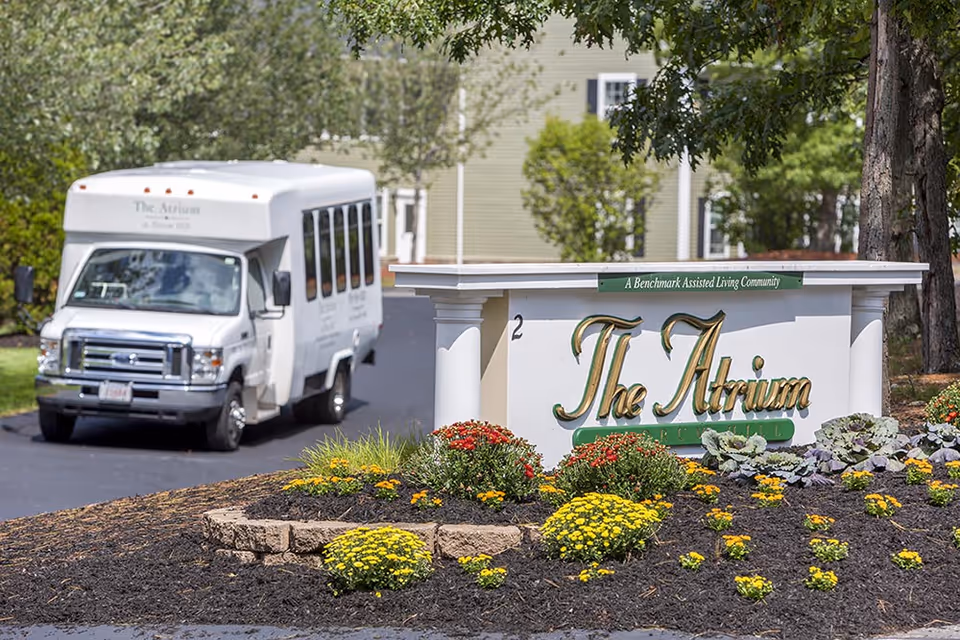 A white shuttle bus with 'The Atrium at Drum Hill' written on it is parked on a driveway near a landscaped flower bed with yellow and red flowers. Next to the flower bed is a white sign with gold and green lettering that reads 'The Atrium' and a smaller green sign above it that says 'A Benchmark Assisted Living Community'. Trees and a building are visible in the background.