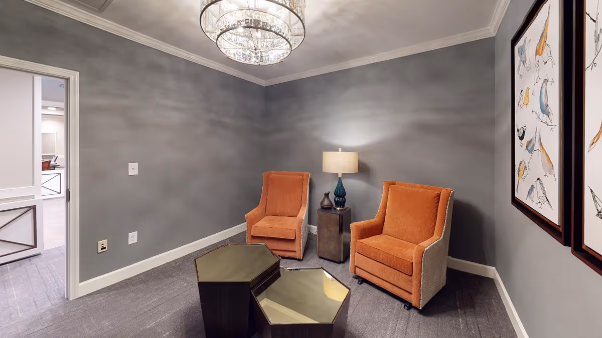 Small interior sitting room with two orange armchairs, a side table with a lamp, a hexagonal coffee table, and framed bird artwork on gray walls.