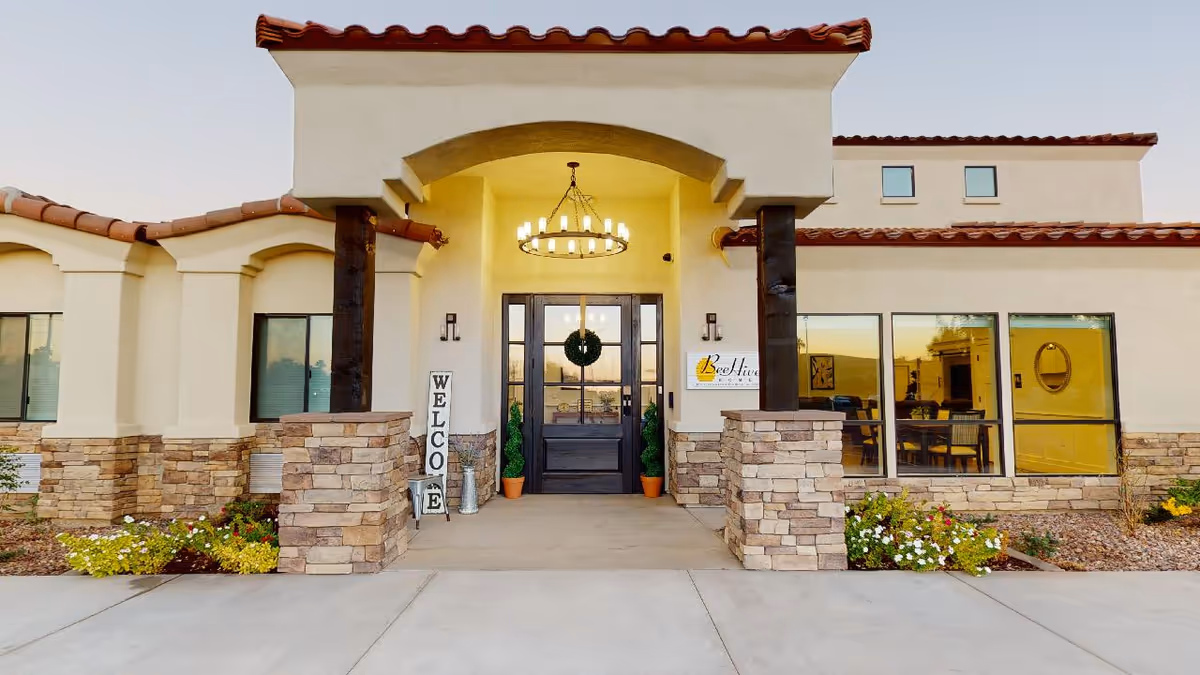 Front entrance of BeeHive Homes Assisted Living facility with a covered porch supported by stone pillars, a chandelier hanging from the ceiling, a wreath on the glass double doors, and a vertical welcome sign on the left side. There are flower beds with colorful flowers on either side of the entrance.
