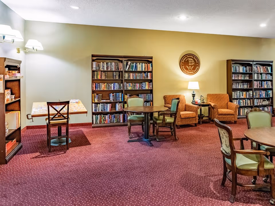 Cozy library-style common room with bookshelves, round tables, chairs, and armchairs under warm lighting.