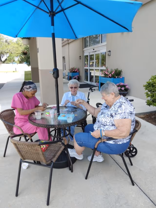 Three women sitting around a round glass table under a large blue umbrella outside a building. Two elderly women and one younger woman in pink scrubs are engaged in an activity involving small items on the table. There are potted flowers near the entrance of the building in the background.