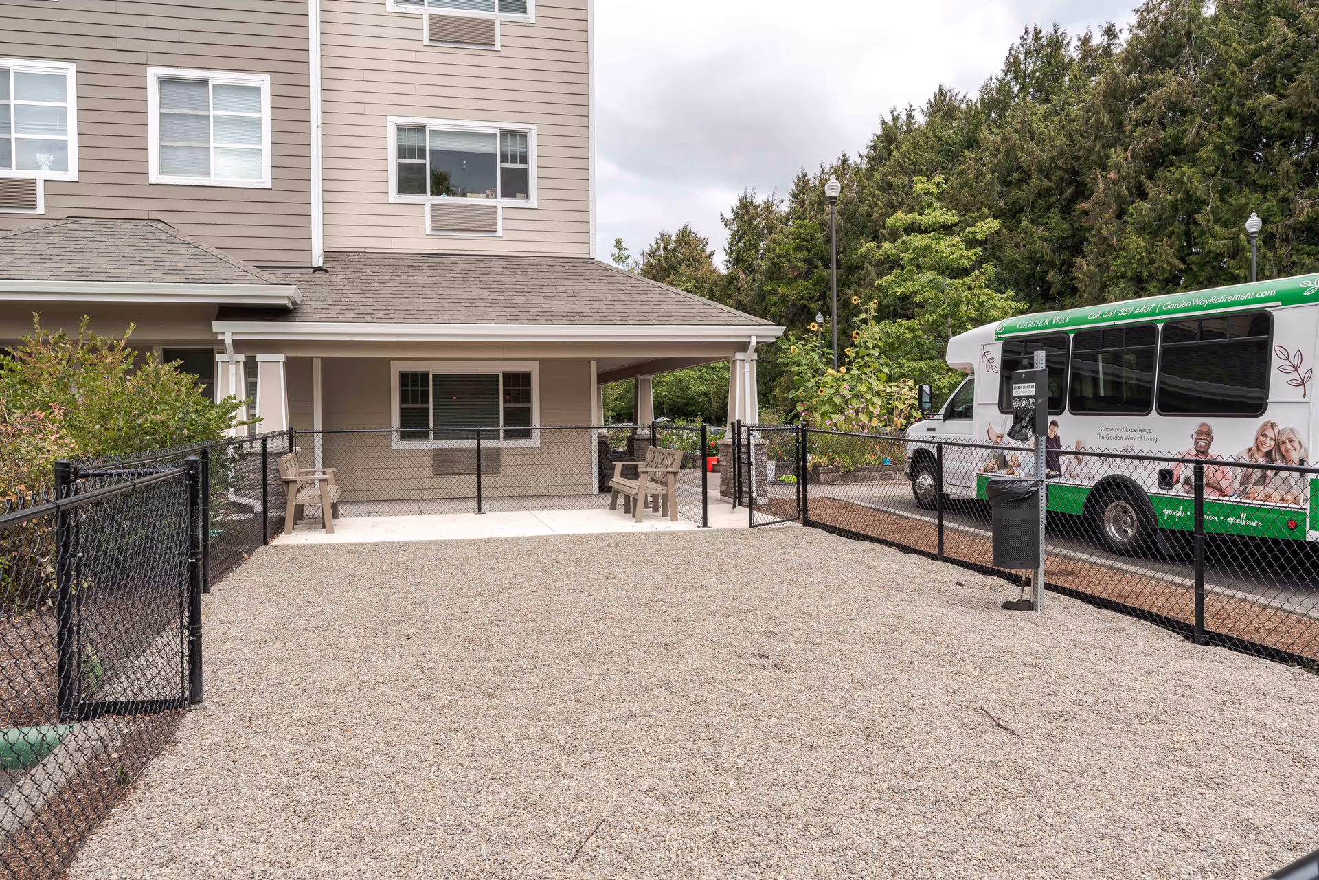 Outdoor area of Garden Way Retirement Community featuring a gravel-covered space enclosed by a black chain-link fence, with a covered patio area furnished with wooden benches. A green and white shuttle bus is parked on the right side near some greenery and trees under a cloudy sky.
