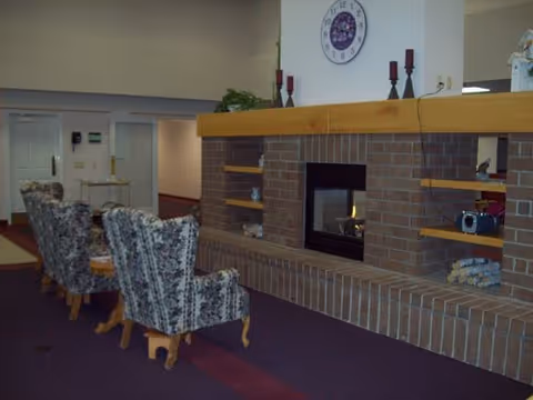 Interior lounge with a brick fireplace, mantle clock and candles, built-in shelves, and three patterned armchairs on purple carpet.