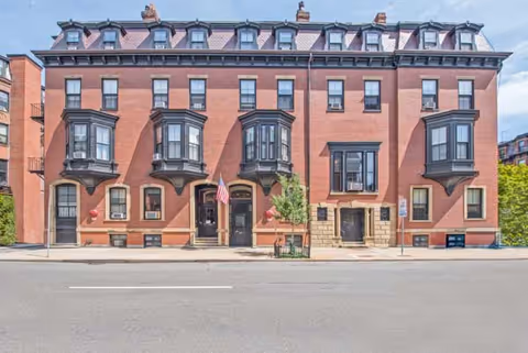 Front exterior view of a large, historic brick building with multiple windows and black bay window structures. The building has a symmetrical design with two main entrances, an American flag, and a small tree planted on the sidewalk in front.