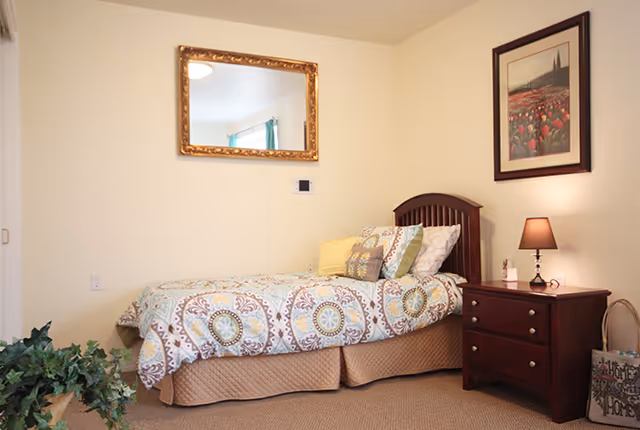 Single bedroom with a patterned twin bed, wooden nightstand and lamp, and framed art and mirror on light-colored walls.