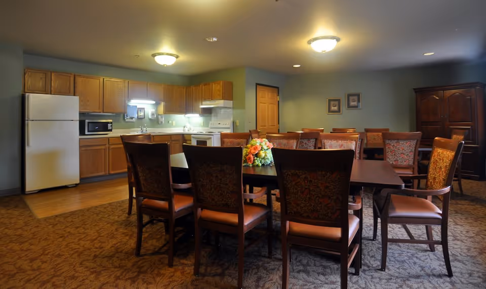 A communal dining area with a large rectangular table surrounded by wooden chairs with patterned upholstery. In the background, there is a kitchen area with wooden cabinets, a white refrigerator, microwave, stove, and sink. The room is softly lit with ceiling lights and has framed pictures on the wall.