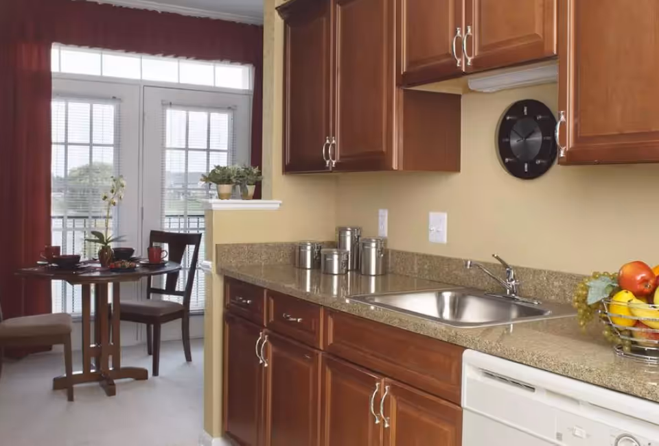 A kitchen with granite countertops and stainless sink, wooden cabinets, a fruit bowl, and a small dining table by glass patio doors.