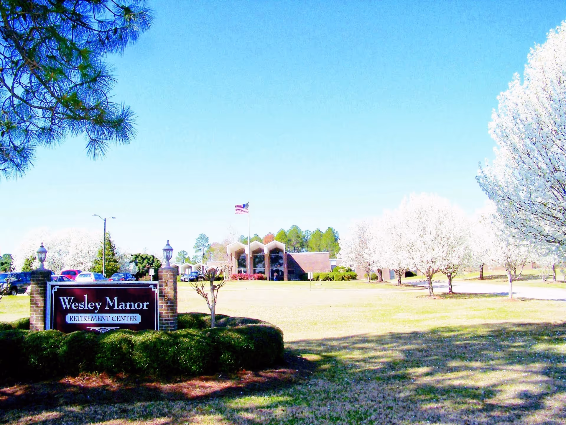 Exterior view of Wesley Manor Retirement Center with a large lawn, blooming white trees, parked cars, and a building with a flagpole displaying the American flag under a clear blue sky.