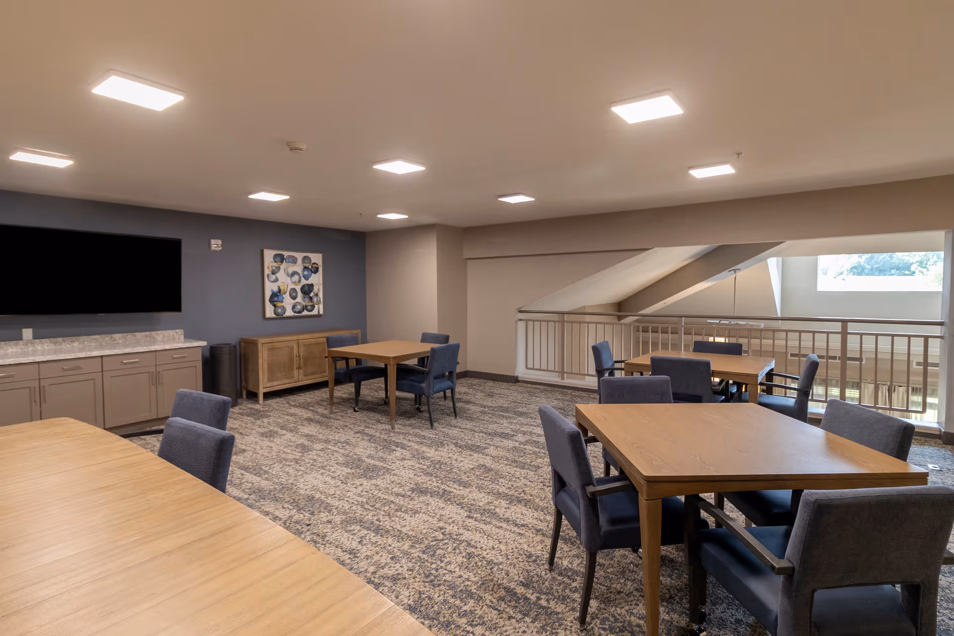 A spacious common area with several wooden tables and blue upholstered chairs arranged around them. The room has a carpeted floor, a large flat-screen TV mounted on a blue accent wall, a wooden sideboard, and a large window letting in natural light. The ceiling has recessed lighting, and there is a railing overlooking a lower level.