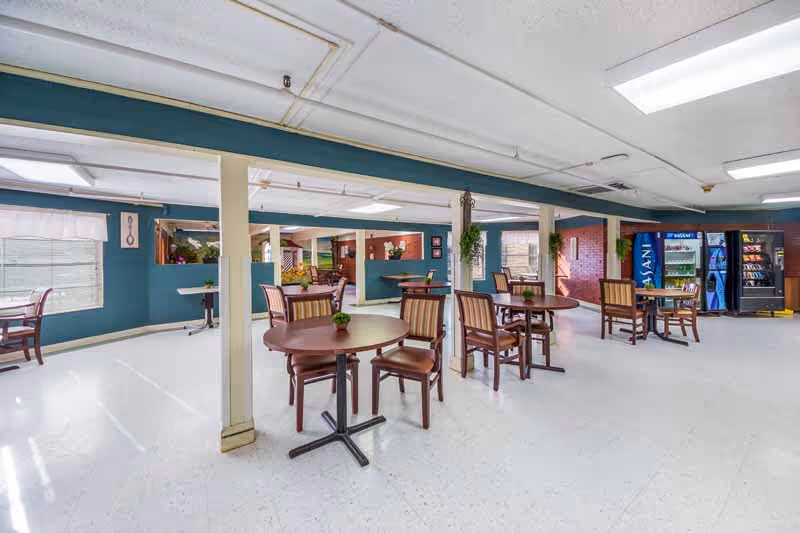 A spacious common area with several round wooden tables and chairs arranged for seating. The walls are painted teal, and there are vending machines against one wall. The room has large windows letting in natural light and some decorative plants on the tables and hanging from the ceiling.