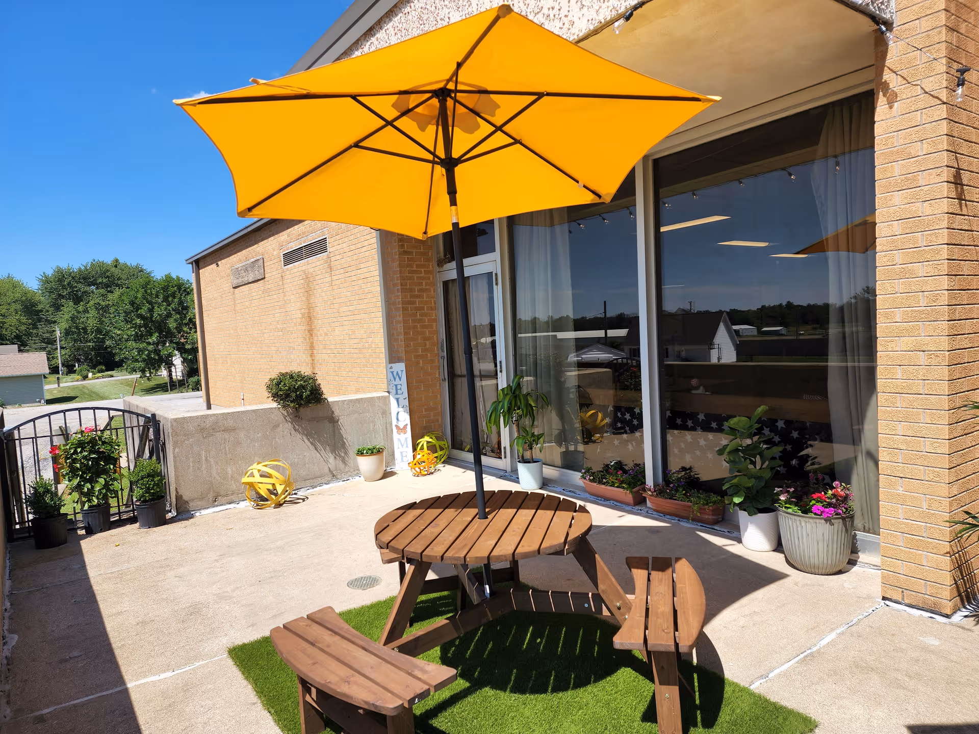 Outdoor patio area with a round wooden picnic table and benches under a large yellow umbrella. The patio is adjacent to a brick building with large windows and several potted plants and flowers arranged along the wall. A small welcome sign is visible near the entrance gate, and the sky is clear and blue.