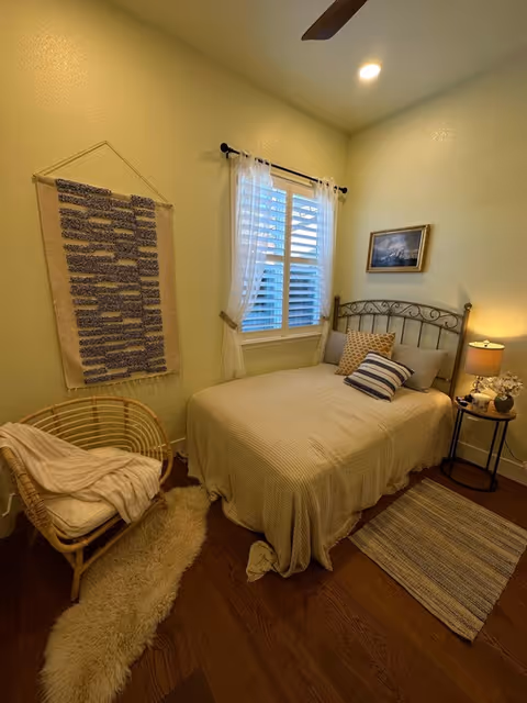 Cozy small bedroom with a bed topped with striped pillows, a nightstand and lamp, a wicker chair, and a window with white curtains.