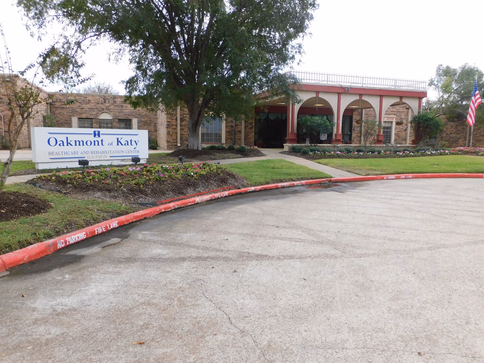 Front exterior view of Oakmont Healthcare and Rehabilitation Center of Katy showing the building entrance with arches, a large tree, landscaped flower beds, a curved driveway with a red curb marked 'No Parking Fire Lane', and an American flag on the right side.