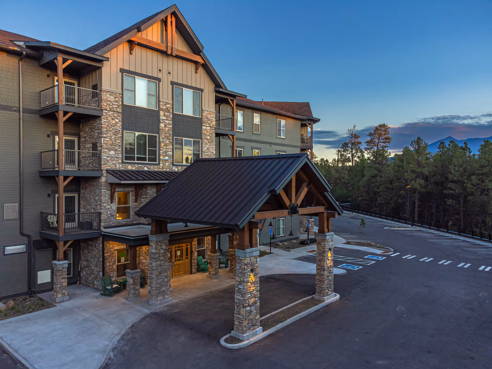 Exterior view of The Bluffs of Flagstaff Senior Living building at dusk, featuring a covered entrance with stone pillars, multiple balconies, and a parking lot with handicap spaces. The building is surrounded by trees and mountains in the background under a clear sky.