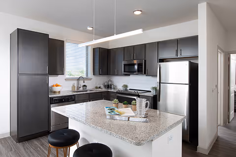 Modern kitchen with dark wood cabinets, stainless steel refrigerator, microwave, and dishwasher. A granite countertop island with two black cushioned stools is in the center, decorated with a tray holding a pitcher, glass, and an open book. A window with blinds is above the sink, and the floor has wood-style flooring.