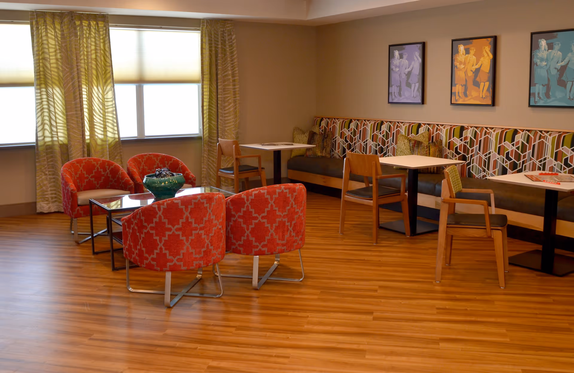 A cozy seating area in a senior living facility with wooden flooring, featuring four red patterned armchairs around a glass coffee table with a decorative bowl, and a built-in bench with geometric patterned upholstery along the wall with three small tables and wooden chairs. Three colorful framed artworks hang on the wall above the bench, and large windows with yellow patterned curtains allow natural light into the room.