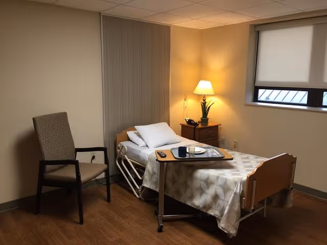 A private nursing room with a single hospital bed, overbed table with dishes, a nightstand and lamp, an armchair, and a window.