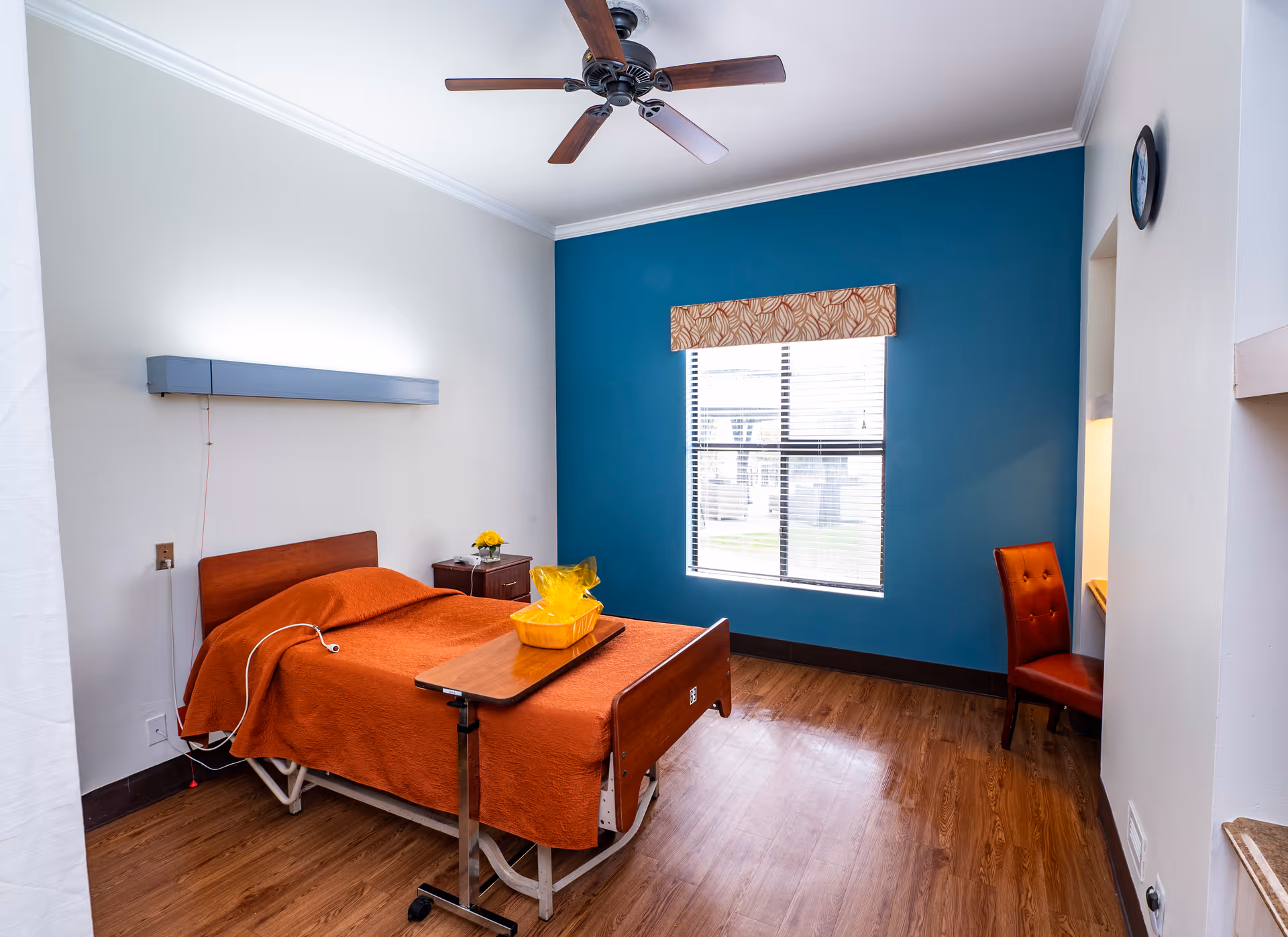 A senior care facility bedroom with a single bed covered in an orange blanket, a small bedside table with a yellow flower vase, a window with blinds and a patterned valance, a ceiling fan, a wooden floor, and a red chair next to a small desk area.