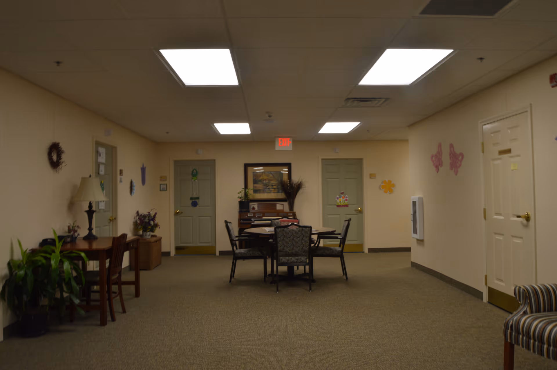 Interior common area of Cedar Cove Assisted Living with a table and four chairs in the center, a piano against the back wall, several closed doors, wall decorations including flowers and butterflies, a lamp on a small table with a chair, and some potted plants.