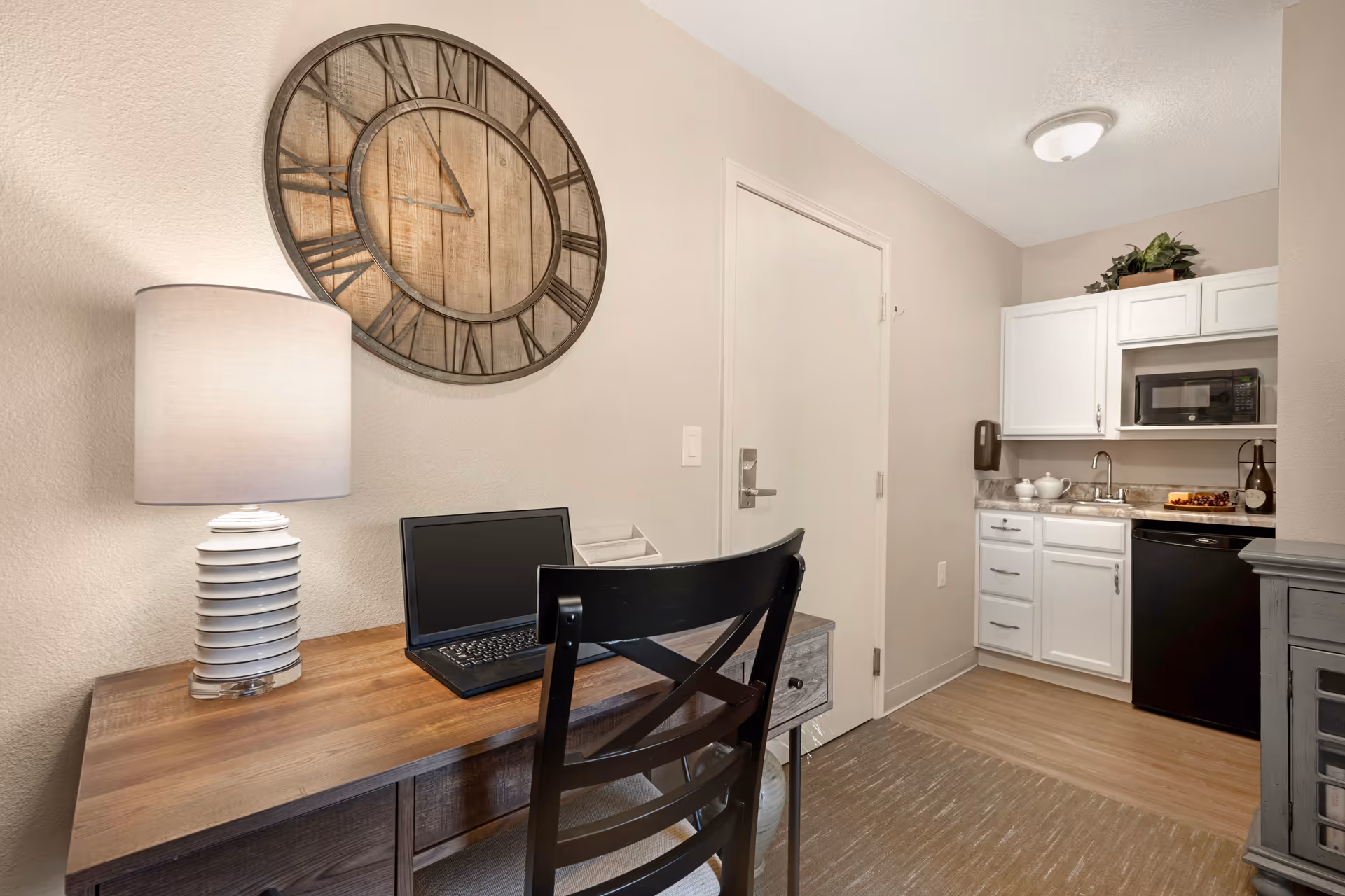 Small apartment-style interior with a desk, lamp and laptop in the foreground and a kitchenette with white cabinets and a microwave in the background.