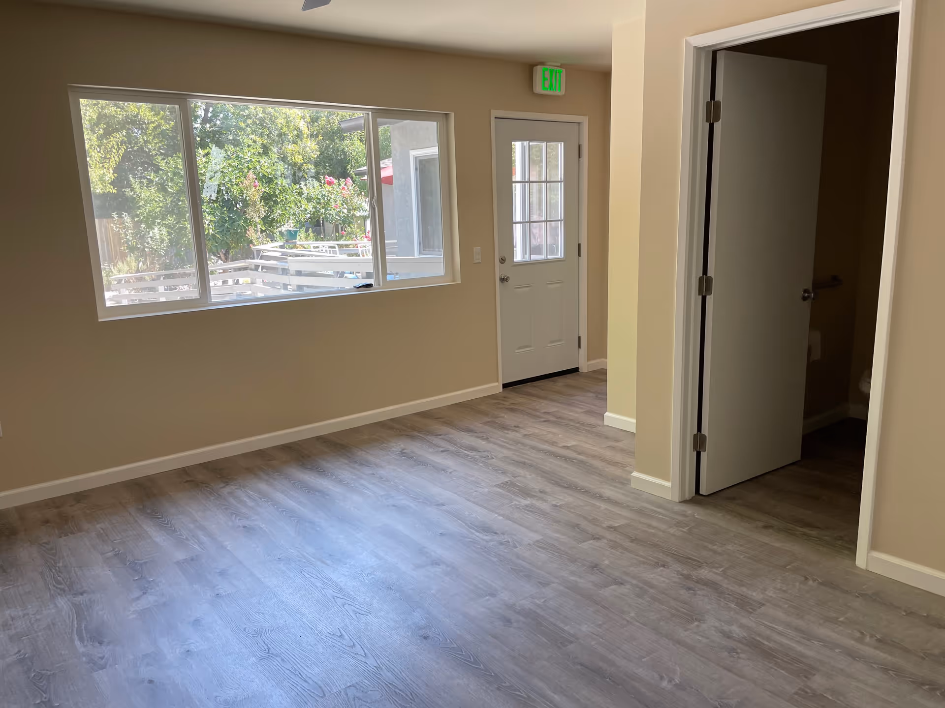 Empty room with light wood flooring, beige walls, a large window showing greenery outside, a white door with a window and an exit sign above it, and an open door leading to a bathroom.