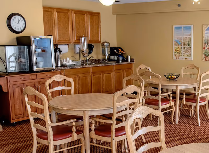 A dining area with round wooden tables and chairs with red cushions. In the background, there is a countertop with wooden cabinets above and below, a small refrigerator, a coffee dispenser, and a water dispenser. The walls are painted light yellow and decorated with two framed pictures. A clock is mounted on the wall above the refrigerator.