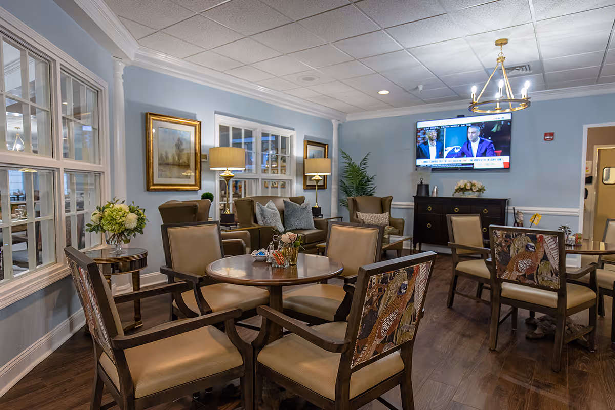 A comfortable communal lounge with round tables and chairs, armchairs, a wall-mounted TV, and soft blue walls.