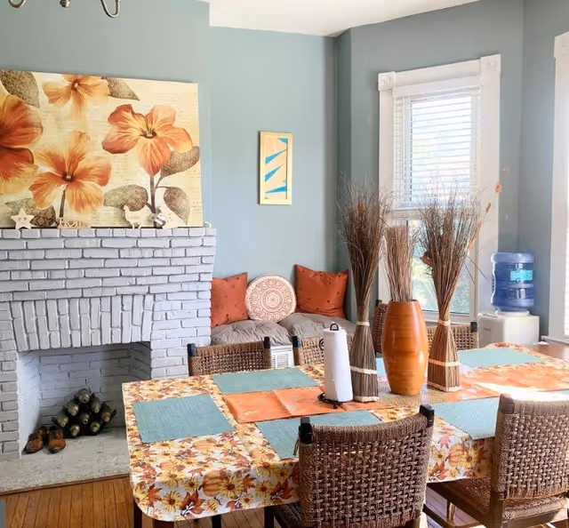 A cozy dining area with a floral tablecloth on the table, four woven chairs, and placemats. The table is decorated with three vases holding dried plants and a paper towel holder. In the background, there is a gray painted brick fireplace with decorative items on the mantel, a cushioned bench with orange and patterned pillows, a window with white blinds, and a water dispenser.