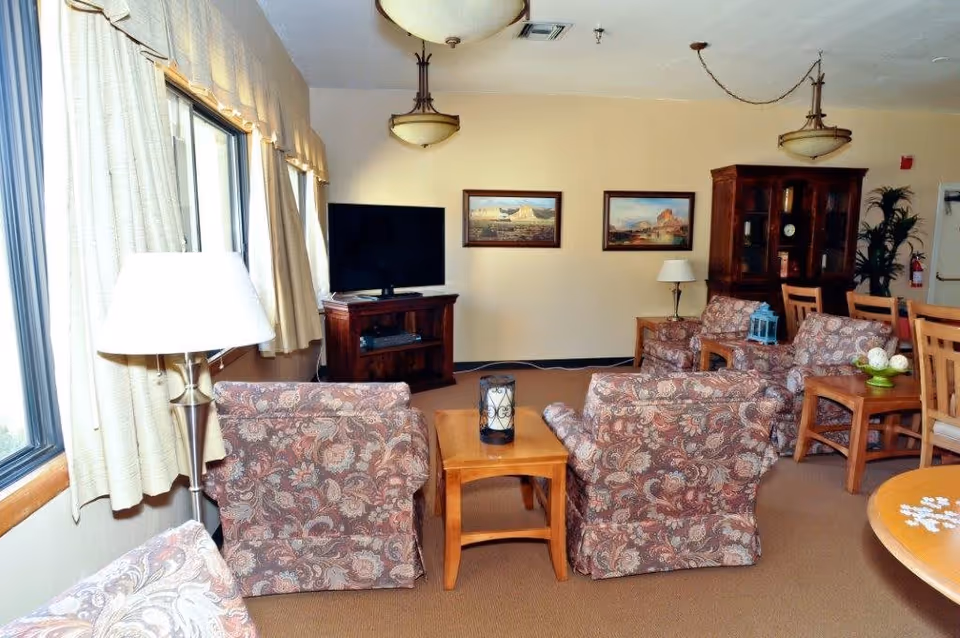 A cozy living room area in an assisted living facility with patterned armchairs arranged around wooden tables. A flat-screen TV is placed on a wooden stand against the wall, which is decorated with two landscape paintings. The room has large windows with beige curtains, ceiling lights, a wooden cabinet, and a dining table with chairs in the background.