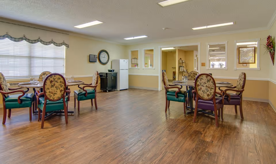 A dining room with two round tables, each surrounded by four upholstered chairs with floral backs and green seats. The room has wooden flooring, beige walls with white trim, a large window with blinds and a valance, a wall clock, a small black cabinet, and a white refrigerator. There are three windows on the far wall looking into another room and a decorative wall hanging with flowers on the right.