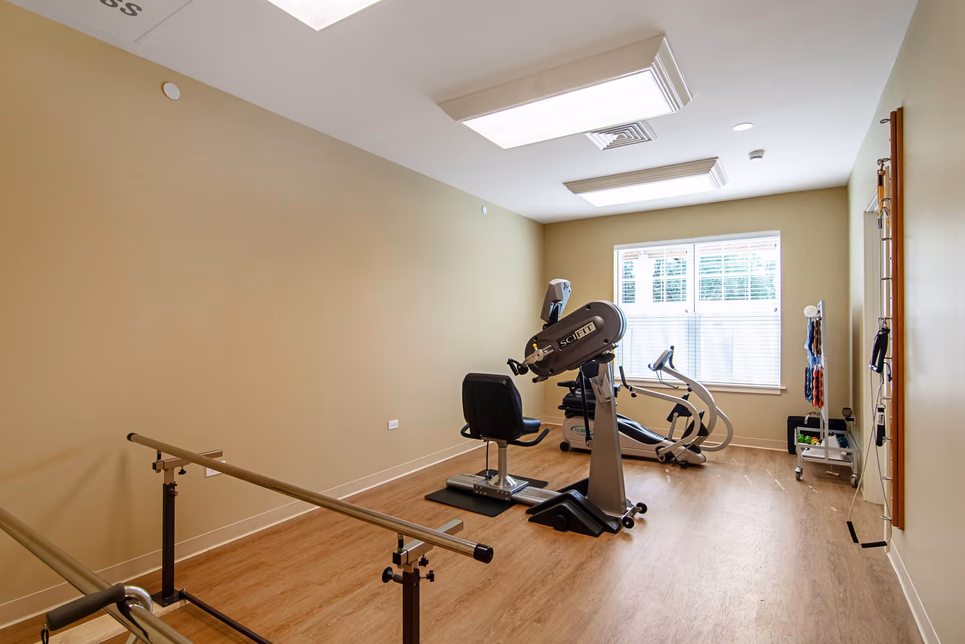A small exercise room with light beige walls and wood flooring. The room contains exercise equipment including a seated arm bike and a recumbent elliptical machine. There are parallel bars on the left side of the room for walking support. A window with white blinds lets in natural light. Various exercise accessories are organized on a rack near the window.
