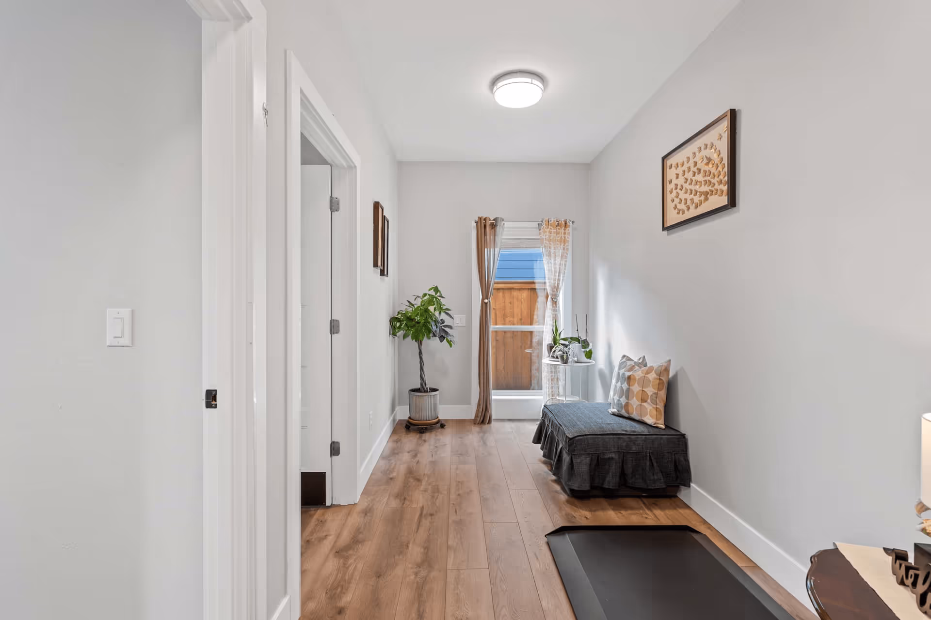 A narrow hallway with light gray walls and wood flooring. On the right side, there is a dark gray cushioned bench with a patterned pillow, a small round table with plants, and a framed artwork on the wall. At the end of the hallway, there is a window with sheer and brown curtains, and a potted plant is placed near the window. On the left side, there is a doorway leading to another room.