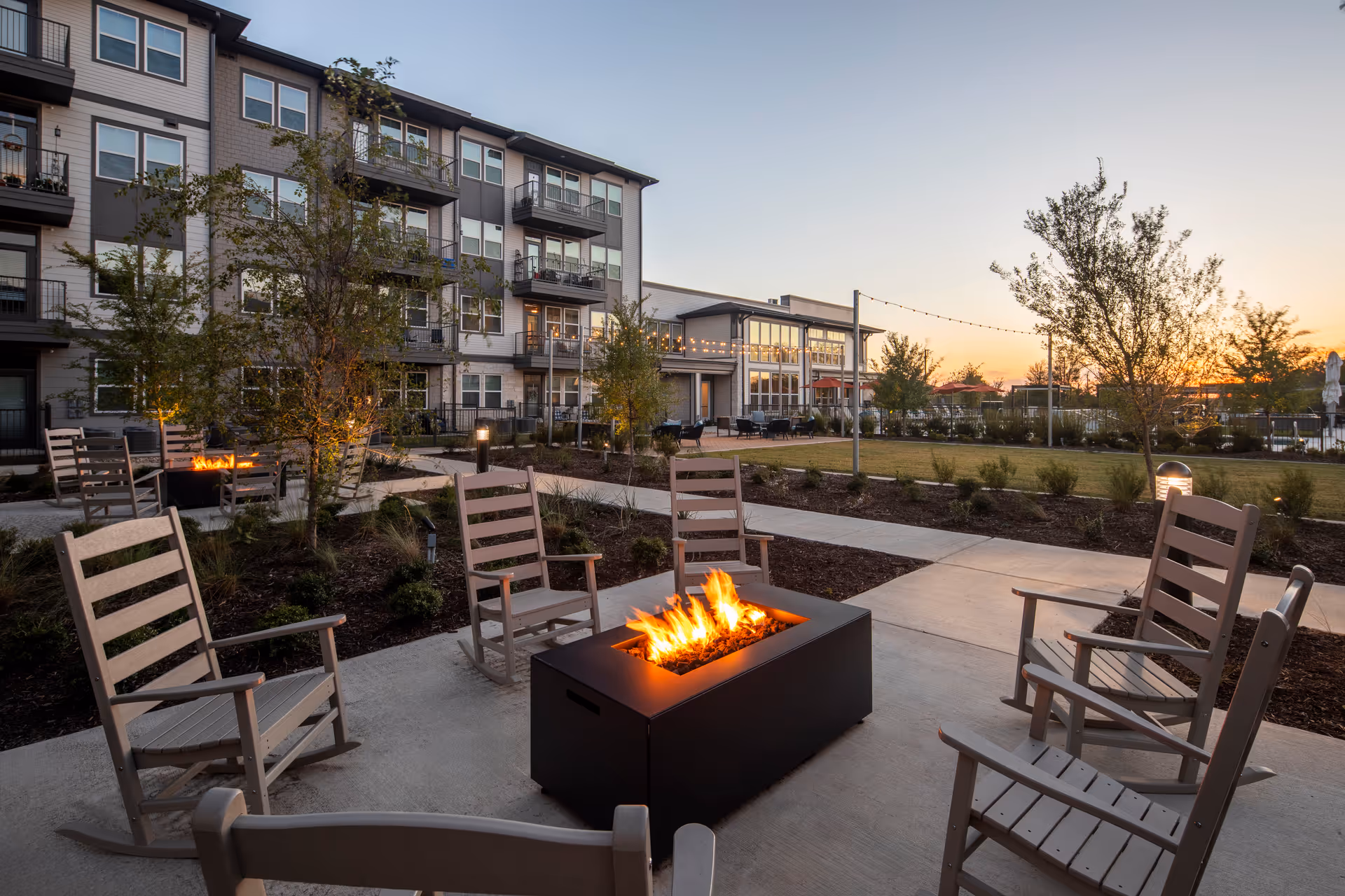 Outdoor seating area at sunset with six wooden rocking chairs arranged around a rectangular fire pit with flames. The area is paved with concrete and surrounded by landscaped plants and trees. In the background, there is a multi-story residential building with balconies and large windows, and string lights are hung across the outdoor space.