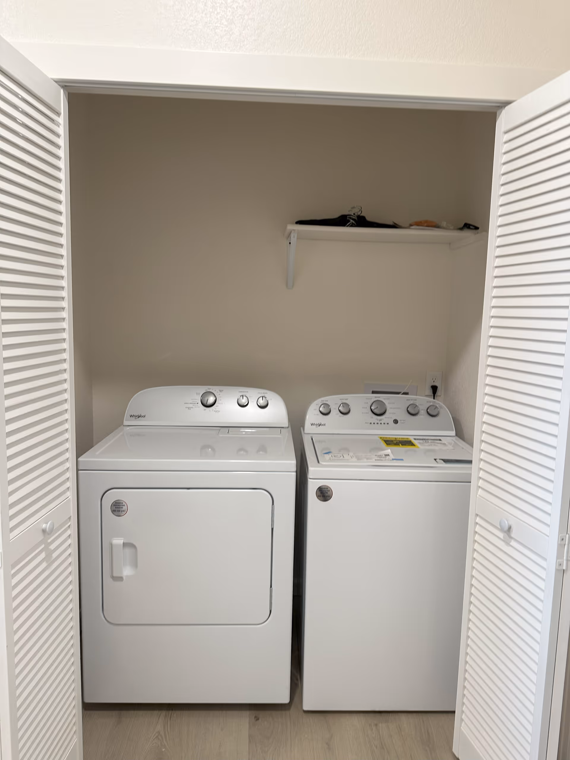 A laundry area with a white Whirlpool dryer on the left and a white Whirlpool washing machine on the right, both placed side by side inside a closet with white louvered doors. Above the machines, there is a white shelf holding a few items against a beige wall.