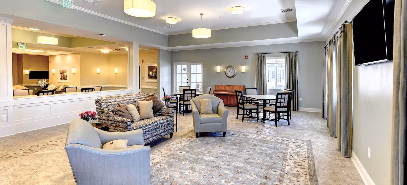 A bright and spacious common area in a senior living facility featuring comfortable seating including a patterned sofa and two armchairs, a large area rug, several tables with chairs, a wall-mounted TV, and large windows with curtains allowing natural light to fill the room.
