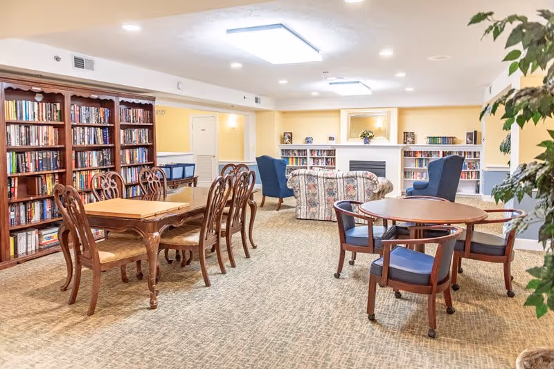 A spacious and well-lit common area in a senior living facility featuring a large wooden table with chairs, a round table with four chairs, bookshelves filled with books, a floral patterned sofa, two blue armchairs, and a white fireplace with decorative items on the mantel. The walls are painted yellow with white trim, and there is a large plant visible on the right side.