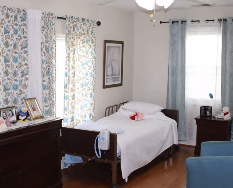 A small, cozy bedroom with a single bed covered in a white blanket and a red stuffed animal on top. The room has wooden flooring, floral patterned curtains on one window, and solid light blue curtains on another. There is a dark wooden dresser with framed photos and a plush toy on it, and a matching nightstand with more framed photos and a white teddy bear. A ceiling fan with lights is visible above.