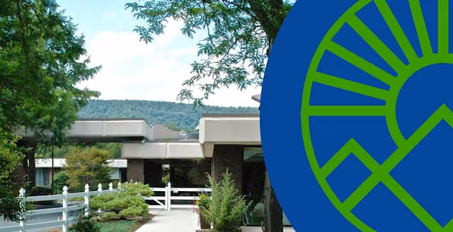 Exterior view of The Terraces at Capitol Village facility showing a walkway with white fencing, greenery including trees and shrubs, and a building with a flat roof in the background under a partly cloudy sky.