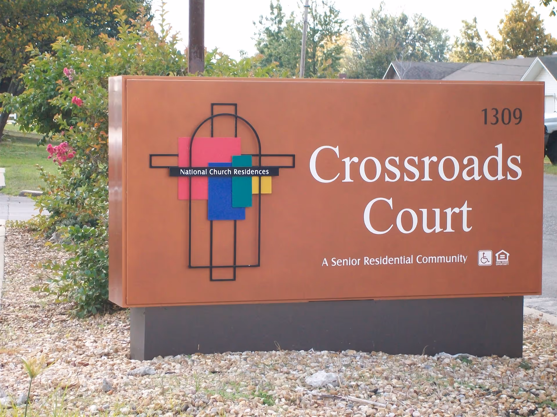 Large brown roadside sign for 'Crossroads Court, A Senior Residential Community' with a colorful National Church Residences logo.