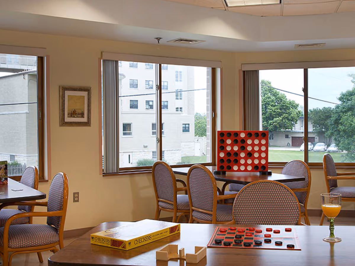A well-lit common area with several tables and chairs arranged near large windows. On one table, there is a large red and black Connect Four game set up, and on another table, a checkers board with pieces in play and a box labeled 'Checkers'. A glass with orange juice is also visible on the table with the checkers game. Outside the windows, buildings and greenery can be seen.