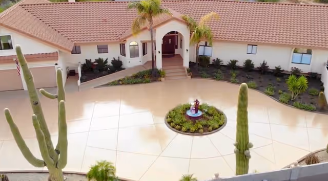 Front exterior view of Estrella Gardens Assisted Living facility with a tiled roof, a circular driveway featuring a small garden with a fountain in the center, and desert landscaping including tall cacti and palm trees.