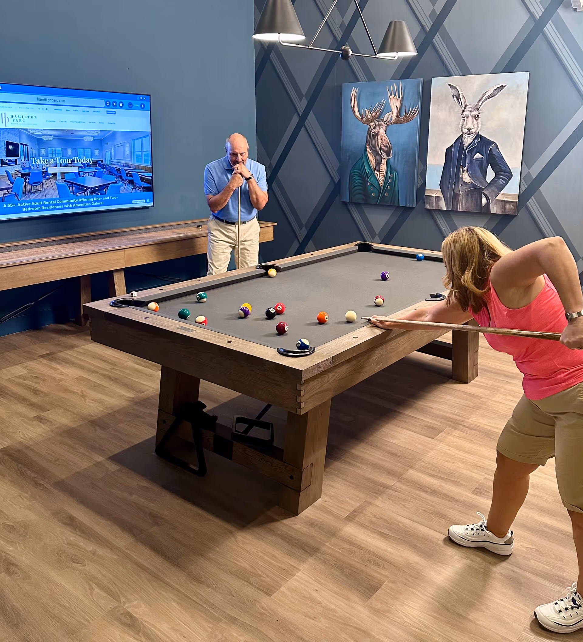Two people playing pool in a game room with a wooden pool table. The man is standing at the far end of the table holding a cue stick, while the woman is leaning over the table aiming a shot. The room has blue walls with two paintings of animals dressed in formal clothing, a moose and a rabbit. A large flat-screen TV mounted on the wall displays the Hamilton Parc website.