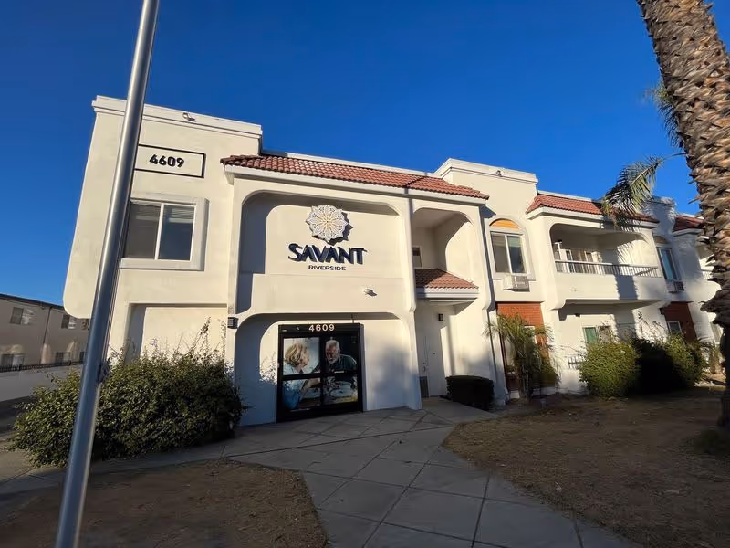 White two-story Mediterranean-style building with 'SAVANT Riverside' signage above a glass entrance marked 4609, palm trees and a clear blue sky.