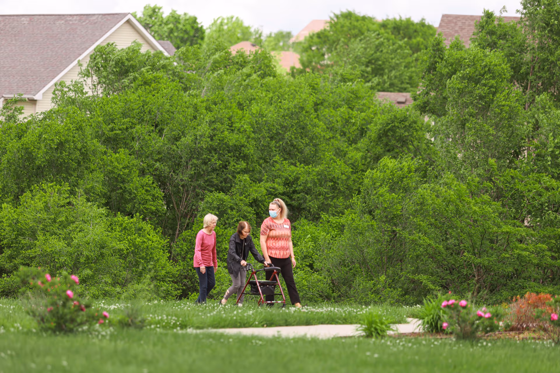 Three people walking along a grassy path near dense trees, with one using a walker and another wearing a face mask.