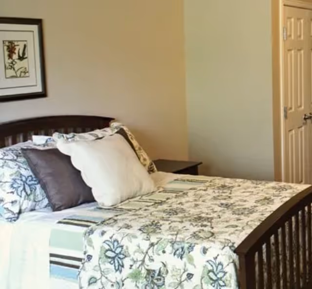A neatly made bed with a floral bedspread and pillows in a simple bedroom with a nightstand and framed wall art.